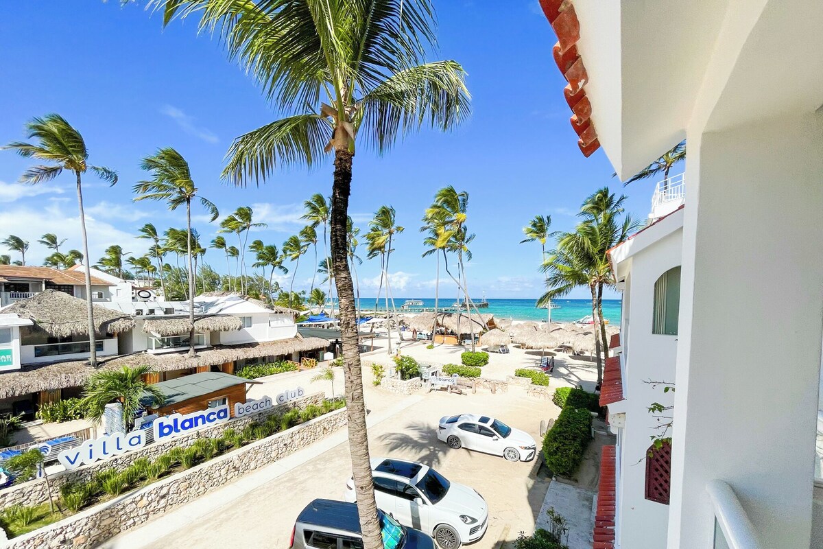 Vista sulla spiaggia con palme, un beach club e una villa chiamata 'Villa Blanca'.
