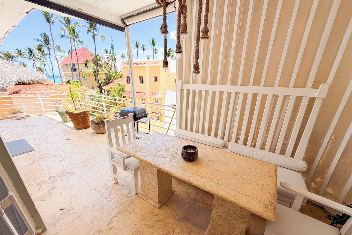 Un balcone con tavolo, sedie, piante in vaso e vista su palme e edifici.