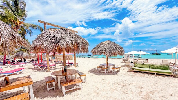Una playa con sombrillas de paja, mesas de madera y sillones de descanso.