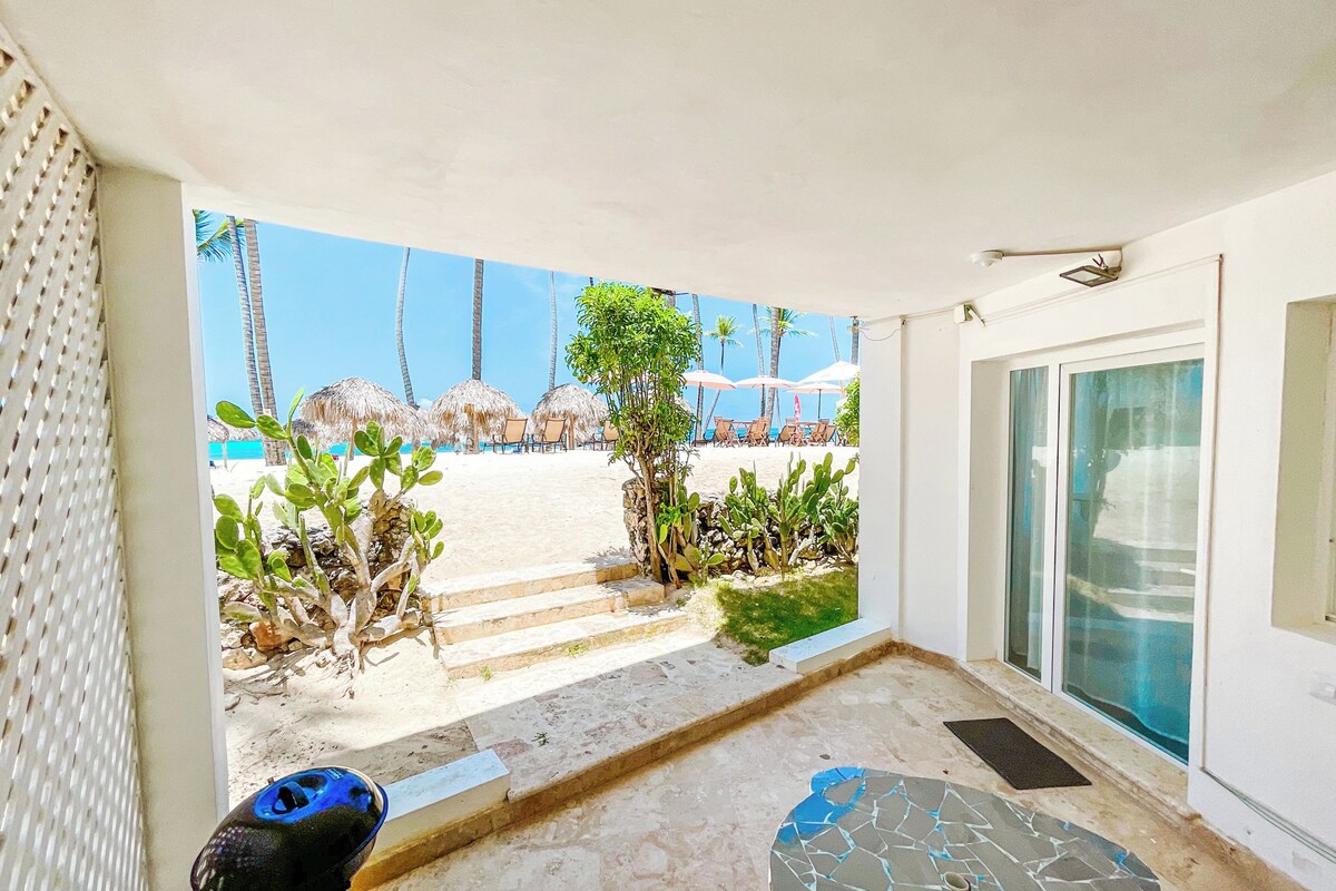 Un'area esterna riparata con patio, barbecue e vista su una spiaggia con ombrelloni e palme.