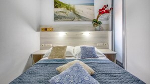 Un dormitorio con cama, mesitas de noche, televisor de pared, una planta y un cuadro con motivos playeros.