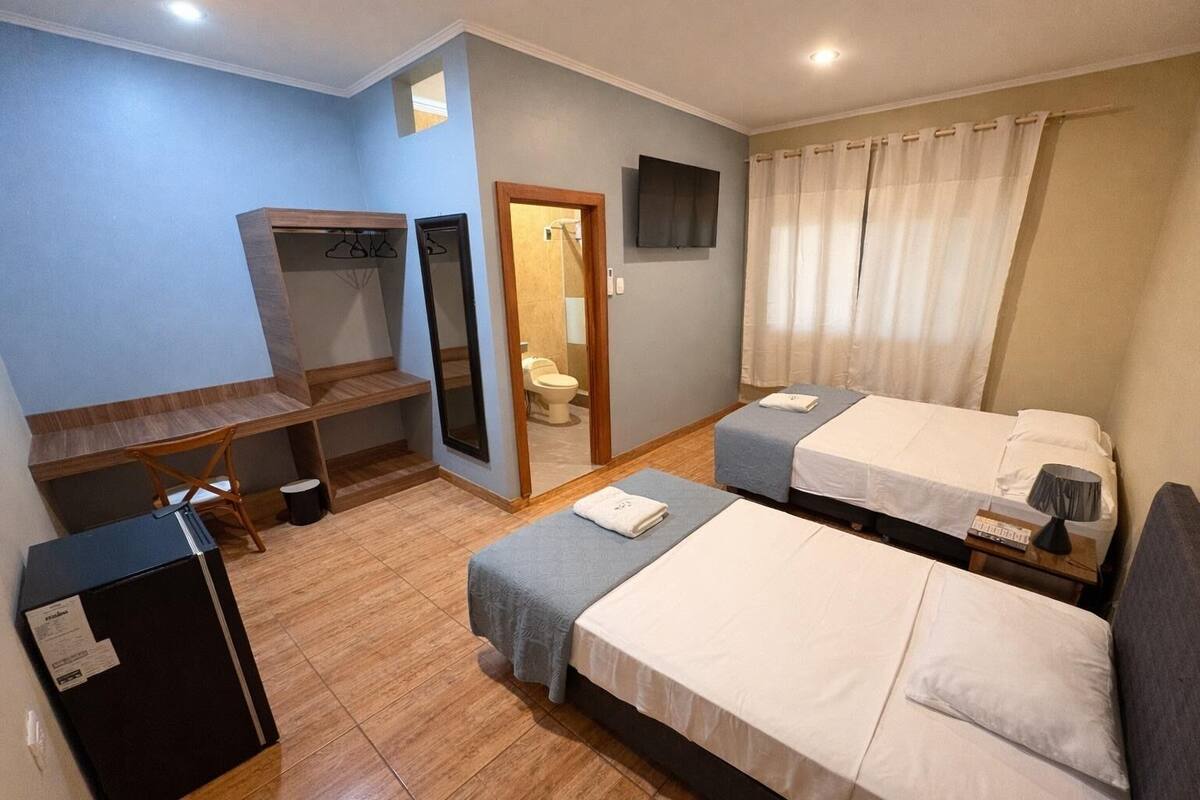 A hotel room with two beds, a desk, a mirror, a TV, and a shower area.