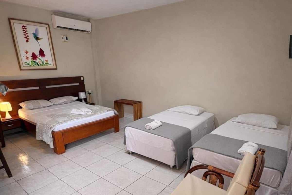 A hotel room with a bed, two single beds, a wooden chair, a small table, and a wall-mounted air conditioner.