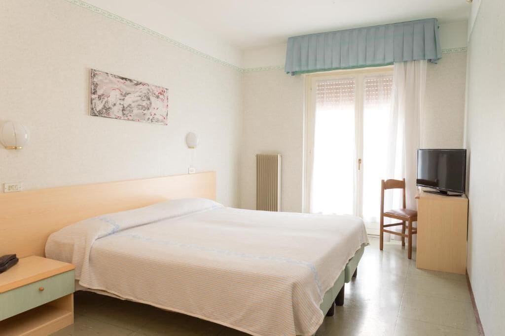 Une chambre d’hôtel avec un lit, une chaise, une télévision, un radiateur et une fenêtre avec des rideaux.