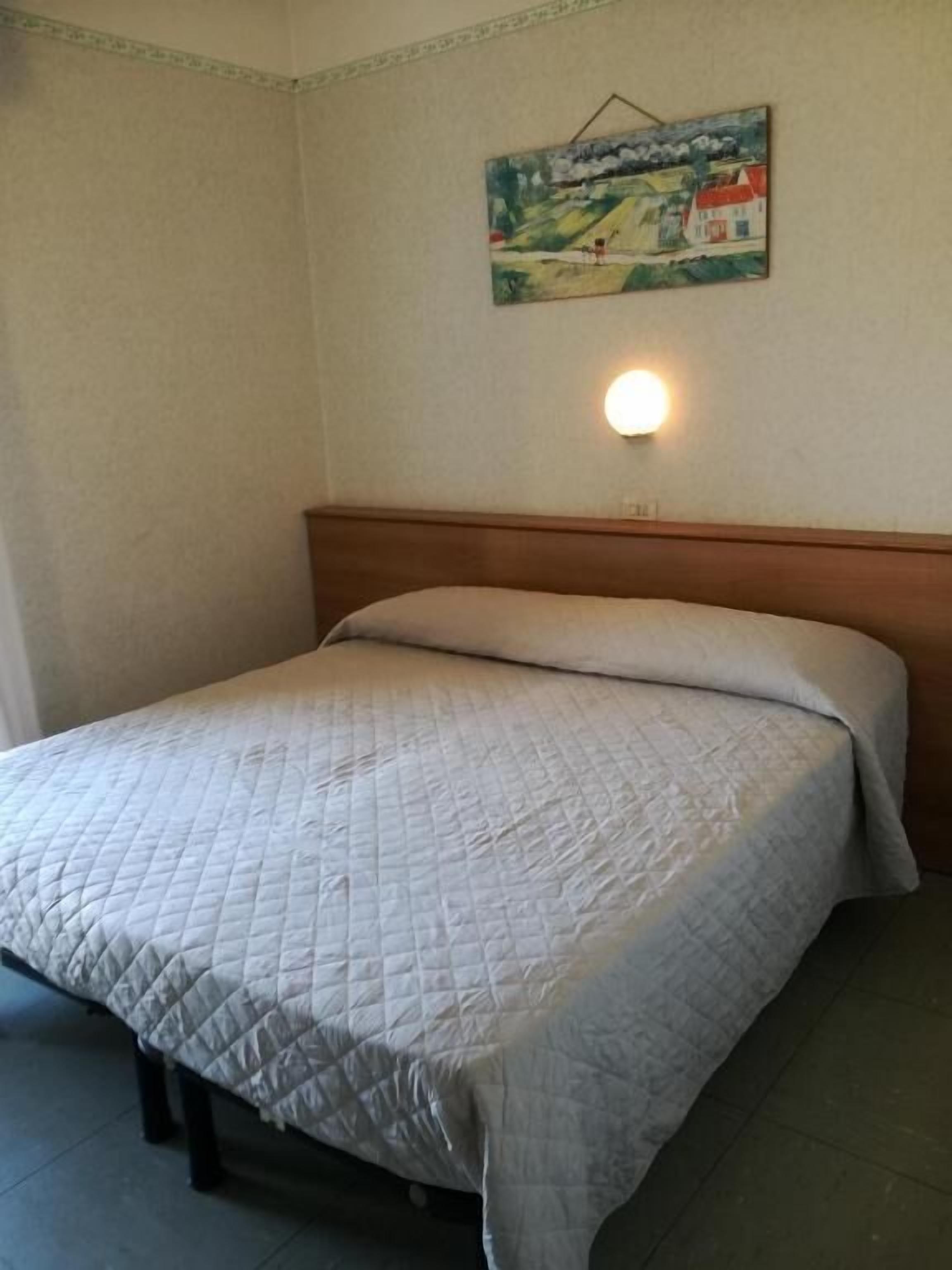 A single bed with a quilted bedspread in a room with a plain wall and a single light fixture.