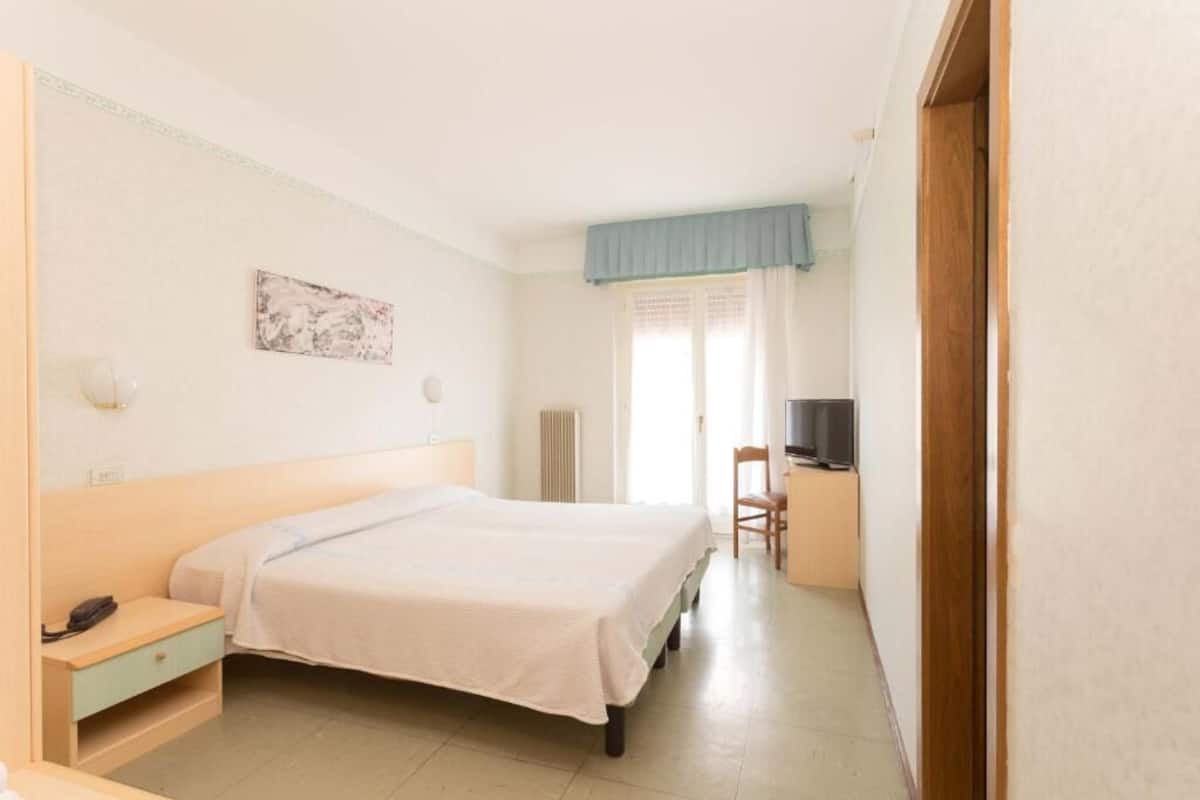 A bedroom with a bed, bedside table, chair, TV, and a window with curtains.