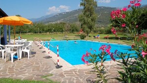 A swimming pool with a stone deck, surrounded by chairs and an umbrella, with mountains in the background. - Aktiv Hotel Eden (Dro)