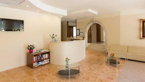 A hotel lobby with a reception desk, a sofa, a bookshelf, and a TV mounted on the wall. - Aktiv Hotel Eden (Dro)