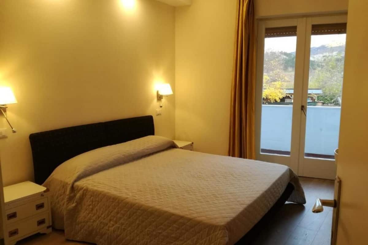 A hotel room with a bed, bedside lamps, a nightstand, and a view of a body of water through a door.