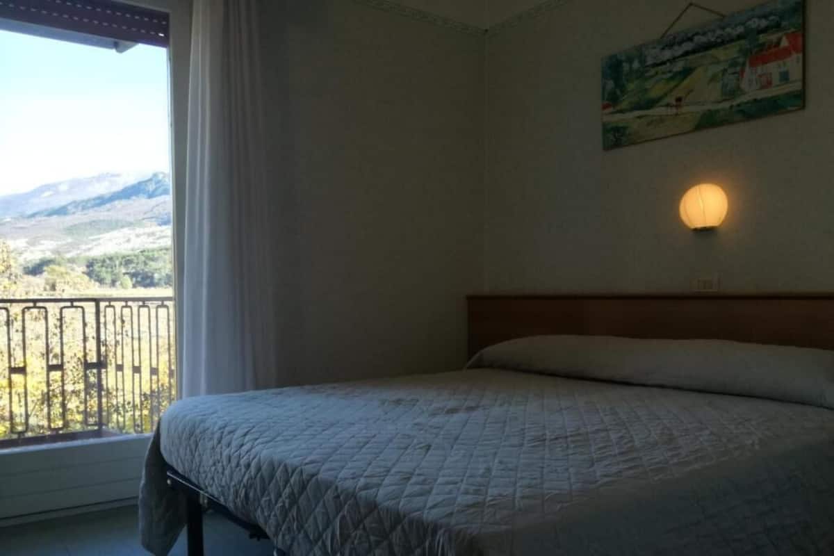 A bedroom with a bed, a window with a view of mountains, a wall-mounted light, and a painting.