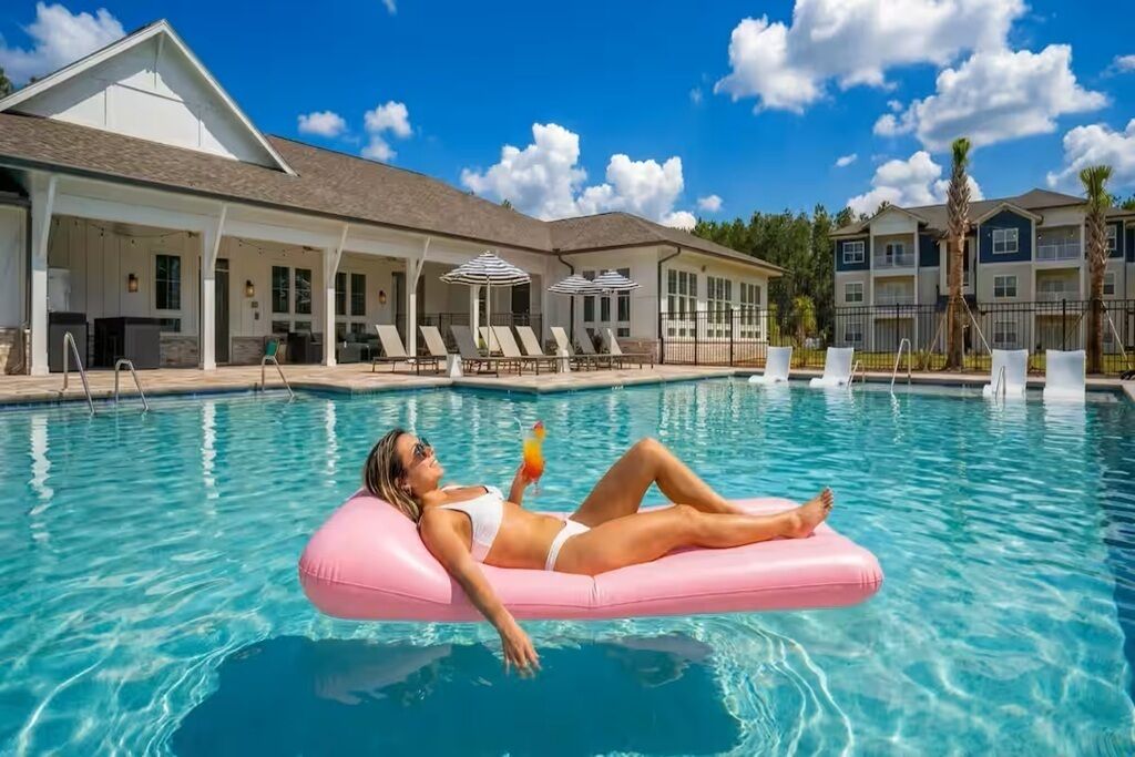 Une piscine avec des chaises longues et des parasols, un bâtiment doté d’un patio couvert et un ciel d’un bleu limpide.