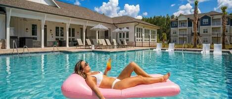 Une piscine avec des chaises longues et des parasols, un bâtiment doté d’un patio couvert et un ciel d’un bleu limpide.