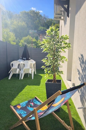Un petit jardin avec une pelouse artificielle, un pot de fleurs, une table pliante avec des chaises et un fauteuil de plage.