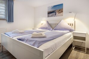 Ein Doppelbett mit weißer und blauer Bettwäsche, zwei Nachttische mit Lampen, ein an der Wand hängendes Bild und ein Holzboden.