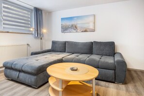 Ein graues Ecksofa mit einem runden Couchtisch, einer Heizung, einem Fenster mit Jalousien und einem Gemälde mit Strandmotiv an der Wand.