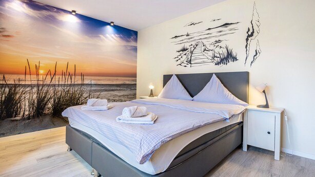 Un dormitorio con una cama grande, una mesita de noche, un mural en la pared con un atardecer playero y un cabecero con diseño.