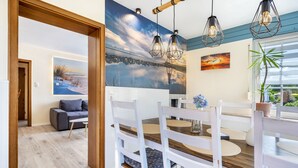 Un comedor con una mesa de madera, sillas blancas y un mural de un atardecer en la pared.