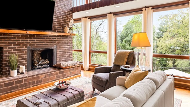 A living room with a brick fireplace, a flat-screen TV, a sofa, and a large window with a view of trees.