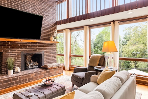 A living room with a brick fireplace, a flat-screen TV, a sofa, and a large window with a view of trees. - Avail Highlands Festival - In-Town Group Getaway (Abingdon)