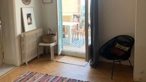 Een kamer met houten vloer, een stoel, een radiator, een lijst met een afbeelding en uitzicht op een terras.