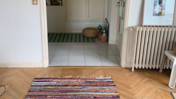 Een kamer met een houten vloer, een gestreept tapijt, een radiator en een deur naar een andere ruimte.