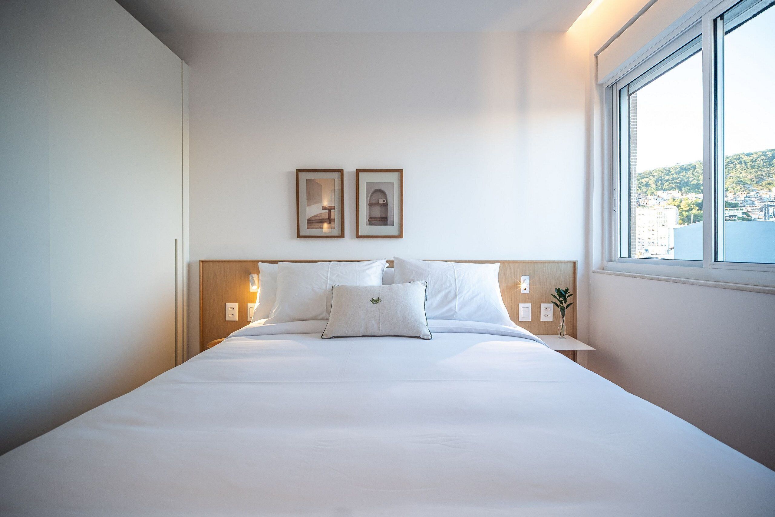 A neatly made bed with white linens, two framed pictures on the wall, and a window with a city view.