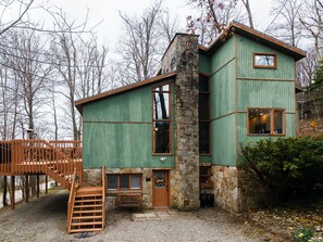 Un chalet rustique doté d’une cheminée en pierre, d’une terrasse en bois et d’un escalier en pierre menant à l’entrée.