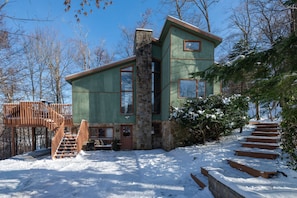 A cabin with a stone chimney, wooden deck, and a staircase leading to a deck area.
