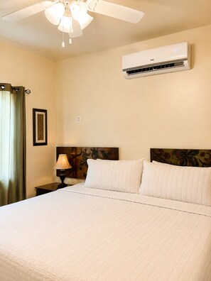 A hotel room with a bed, an air conditioner, a ceiling fan, a nightstand with a lamp, and a framed picture on the wall. - Tranquil North Ambergris Caye Retreat - 3BR Home - Sleeps 8 (San Pedro)