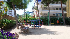 Ein Spielplatz mit Schaukeln und einer Bank, umgeben von Bäumen und einem Gebäude im Hintergrund.