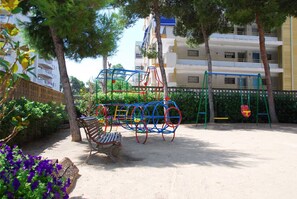 A playground with swings and a bench, surrounded by trees and a building in the background. - Apartment with pool near Cala Capellans (Salou)