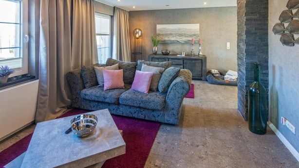 A living room with a sofa, a coffee table, a fireplace, and a large window. - Vacation apartment Meeresrauschen (Hagermarsch)