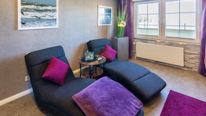 A room with two dark sofas, a small table with a glass, a purple throw, a tall plant, and a window with blinds. - Vacation apartment Meeresrauschen (Hagermarsch)