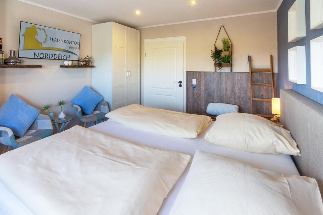 Ein Hotelzimmer mit zwei Betten, einem Sofa, einem kleinen Tisch und einem Sessel. Eine Tür, ein an der Wand befestigtes Regal mit Pflanzen und ein Schild sind ebenfalls vorhanden.