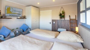 A hotel room with two beds, a sofa, a small table, and a chair. There is a door, a wall-mounted shelf with plants, and a sign on the wall. - Vacation apartment Treibholz | Quiet North Sea vacation Hilgenriedersiel (Hagermarsch)