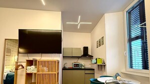 Ett kompakt bostadsutrymme med ett litet kök, en matplats med stolar, en TV och ett fönster med persienner.