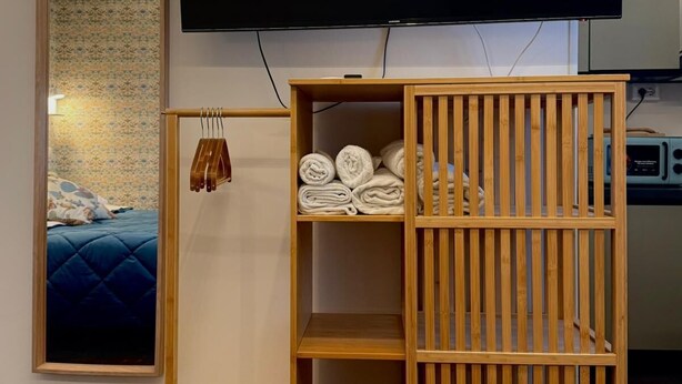Ett rum med en stor, platt-TV som är monterad på väggen, ett träskåp med hyllor, en spegel som reflekterar en säng och ett litet träbord.