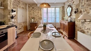Une pièce aux murs de pierre, avec une longue table à manger dressée pour quatre personnes, un luminaire suspendu et une fenêtre avec des rideaux.
