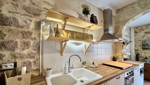 Une cuisine avec un mur en pierre, un plan de travail en bois, des placards blancs, un évier et une étagère suspendue ornée d’une enseigne.