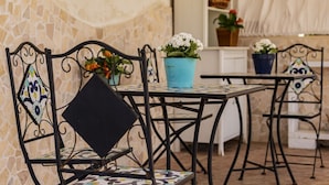 Buiten zitplaatsen met metalen stoelen en tafels, potplanten en een stenen muur.