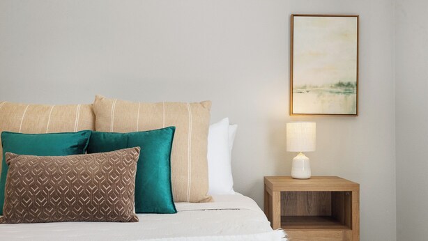 Una cama con varios almohadones, una mesita de noche con lámpara y un cuadro enmarcado en la pared.
