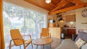 Un espacio de sala de estar con un sofá, dos sillones, una mesa de madera, una cocina con armarios y electrodomésticos, y un ventanal con cortinas.
