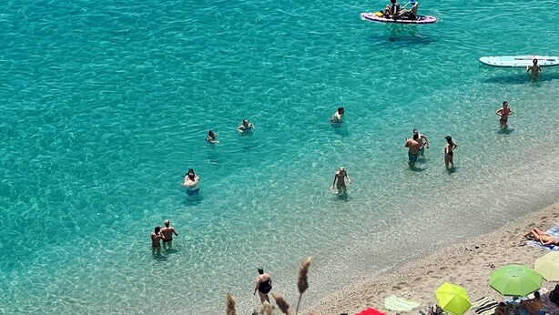 Una spiaggia con acque cristalline turchesi, persone che nuotano e fanno paddleboarding, e ombrelloni colorati sulla sabbia.