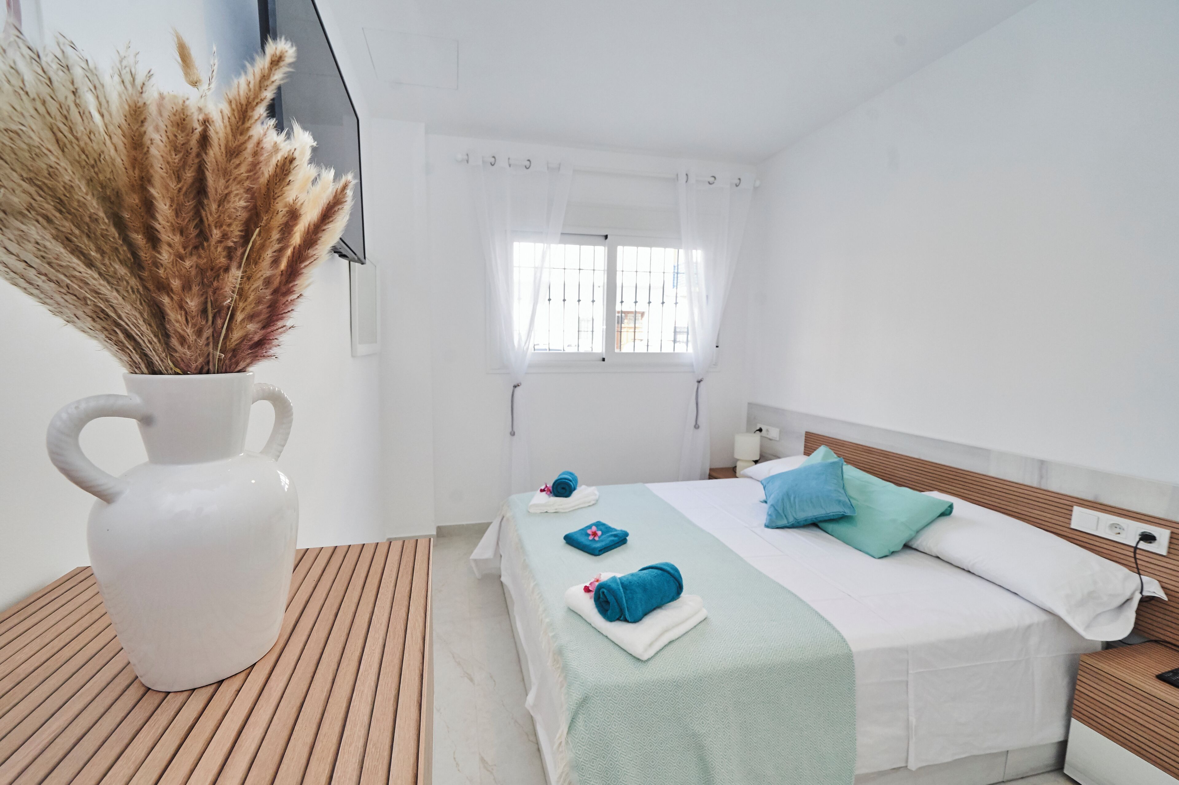 Une chambre d’hôtel avec un lit, une table de chevet, une commode, une fenêtre avec des rideaux, un vase blanc contenant des plumes d’herbe des pampas et un plancher en bois.