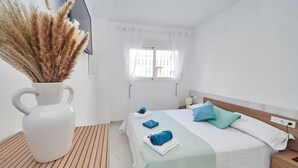 Una camera d'albergo con un letto, un comodino, tende a una finestra, un vaso bianco con pampas e un pavimento in legno.