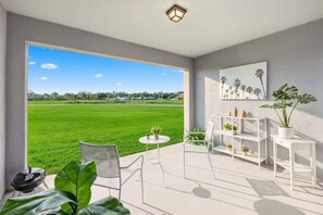 A modern room with a large window offering a view of a green field, a white chair, a small round table, and a white shelving unit with plants. - Orlando Family Retreat Near Disney Magic (Davenport)