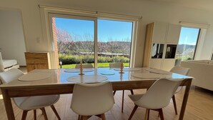 Een eetgedeelte met een houten tafel en witte stoelen, grote ramen met uitzicht op bomen en heuvels, en een witte bank op de achtergrond.