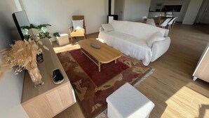 Een moderne woonkamer met een bank, salontafel en stoelen, een keuken op de achtergrond en een kleine tafel met een bloemenvaas.