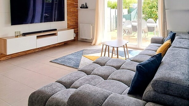 A modern living room with a large flat-screen TV, a grey sectional sofa, a dining table with chairs, and a sliding glass door leading to an outdoor area. - Le Refuge Balnéaire - À 3km de la plage (Pornichet)