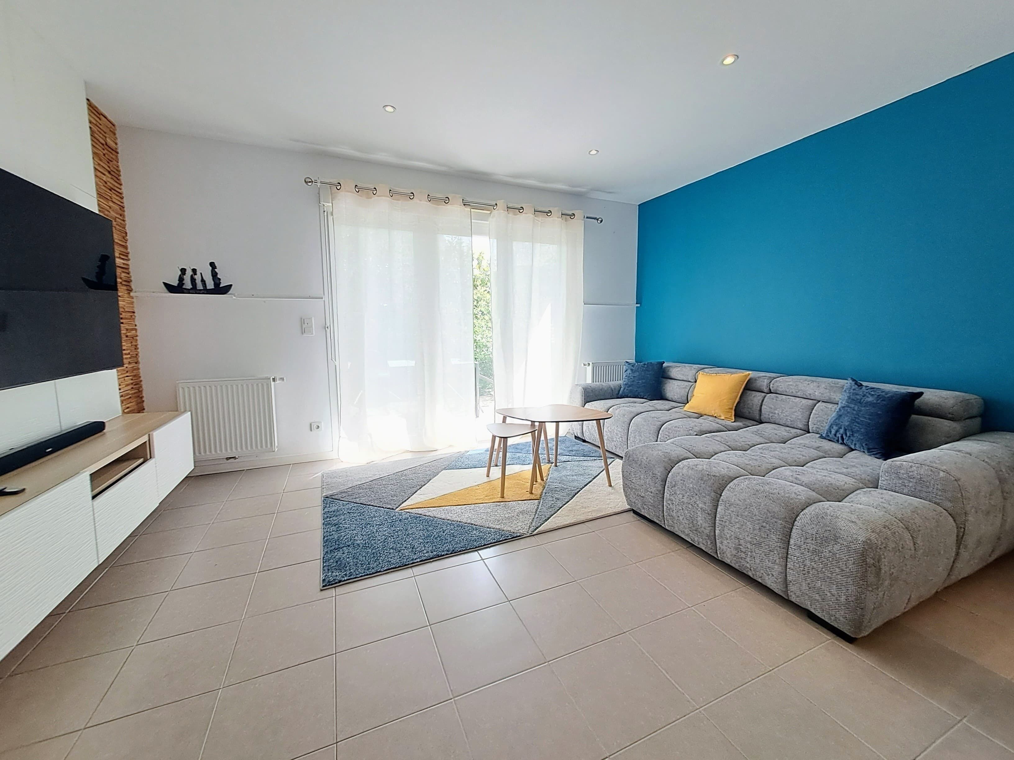 A modern living room with a grey sofa, a small coffee table, a flat-screen TV, and a large window with sheer curtains.